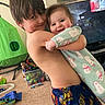 Aurora is registered to the contest to win money with this photo: child, baby, hug, smile, living_room, toys, play_tent, pajamas, television, carpet, wall_art, clothing, person, indoor, brother_and_sister, family, happy, cute, home, playtime