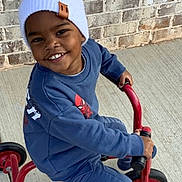 Benson joined the competition — help win amazing prizes! child, smiling, tricycle, blue_clothing, white_beanie, outdoor, concrete, brick_wall, happy, toy, play, person, footwear, casual, young, kid, transportation, headwear, vehicle, daylight