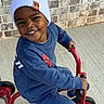child, smiling, tricycle, blue_clothing, white_beanie, outdoor, concrete, brick_wall, happy, toy, play, person, footwear, casual, young, kid, transportation, headwear, vehicle, daylight