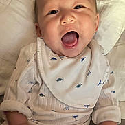 Su-Jin joined the competition — help win amazing prizes! baby, bib, child, close_up, cozy, cute, dinosaur_pattern, happy, indoors, infant, lying_down, open_mouth, pajamas, person, portrait, skin, smiling, soft_surface, white_clothing, young