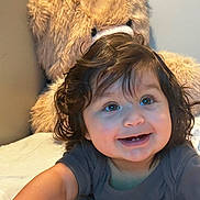 Ashley is registered to the contest to win money with this photo: baby, child, smile, curly_hair, teddy_bear, plush_toy, bed, indoor, cute, happy, infant, person, soft_toy, face, young_child, cuddly, portrait, lying_down, blue_eyes, casual_clothing