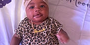 Honestii is registered to the contest to win money with this photo: baby, infant, headband, bow, leopard_print, onesie, necklace, blanket, text, lying_down, cute, child, jewelry, portrait, indoors, person, skin, face, expression, young