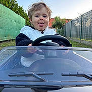 Louca participe au concours pour gagner de l'argent avec cette photo : child, toddler, toy_car, steering_wheel, outdoor, greenery, fence, tongue_out, happy, playful, casual_clothing, daylight, young_child, portrait, fun, smiling, hair, person, nature, backyard
