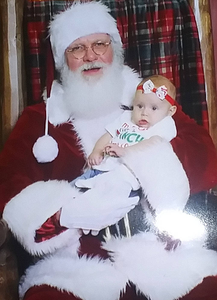 Journee is registered to the contest to win money with this photo: baby_toddler_clothing, beard, christmas, christmas_eve, comfort, curtain, event, face, facial_expression, facial_hair, hat, headwear, holiday, human_body, lap, person, red, santa_claus, sleeve, standing