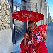 Ellote participe au concours pour gagner de l'argent avec cette photo : child, girl, red_dress, traditional_clothing, parasol, fan, street, sidewalk, stone_wall, building, smile, braided_hair, outdoor, daytime, cultural, portrait, fashion, urban, colorful, accessory