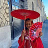 child, girl, red_dress, traditional_clothing, parasol, fan, street, sidewalk, stone_wall, building, smile, braided_hair, outdoor, daytime, cultural, portrait, fashion, urban, colorful, accessory