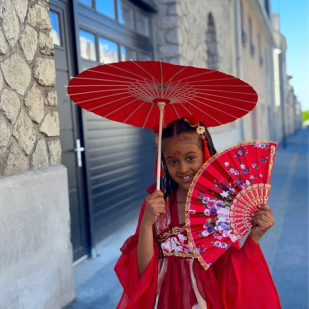 Ellote participe au concours pour gagner de l'argent avec cette photo : child, girl, red_dress, traditional_clothing, parasol, fan, street, sidewalk, stone_wall, building, smile, braided_hair, outdoor, daytime, cultural, portrait, fashion, urban, colorful, accessory