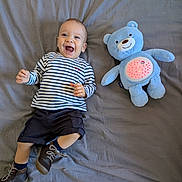 Fatah Juba participe au concours pour gagner de l'argent avec cette photo : baby, child, striped_shirt, shorts, brown_shoes, blue_plush_bear, toy, smiling, lying_down, gray_bedspread, happy, cute, indoor, soft_toy, fabric, footwear, infant, playful, toddler, portrait