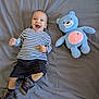 baby, child, striped_shirt, shorts, brown_shoes, blue_plush_bear, toy, smiling, lying_down, gray_bedspread, happy, cute, indoor, soft_toy, fabric, footwear, infant, playful, toddler, portrait