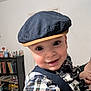 baby, child, face, cap, hat, plaid_shirt, suspenders, smiling, indoor, person, hand, bookshelf, books, furniture, clothing, portrait, cute, happy, headwear, young
