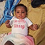 baby, infant, child, clothing, shirt, skirt, blanket, text, tongue_out, cheeky, lying_down, fabric, colorful, cute, person, indoors, playful, expression, face, young_child