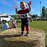 Tiago participe au concours pour gagner de l'argent avec cette photo : toddler, child, hay_bale, outdoor, sunny, grass, field, fence, smiling, people, clothing, trees, tent, daylight, blue_sky, shoes, joyful, park, casual, festival