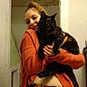 animal, black_cat, casual_clothing, cat, cozy, cute, doorway, feline, friendly, happy, holding, home, indoor, orange_sweater, person, pet, portrait, smiling, tortoiseshell_cat, wall