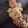 baby, child, plush_toy, unicorn, couch, sofa, indoor, clothing, pants, yellow_top, hair_clip, cute, portrait, infant, sitting, animal_print, soft_toy, looking_at_camera, cozy, relaxed