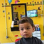 child, boy, haircut, barbershop, mirror, yellow_wall, video_game_decor, red_chair, black_cloth, reflection, television, shelf, bottle, cabinet, hair_style, expression, indoor, person, furniture, wall_art