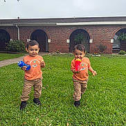 Mateo And Matias joined the competition — help win amazing prizes! child, toddler, grass, lawn, orange_shirt, khaki_pants, sippy_cup, snack, brick_building, arched_doorways, greenery, outdoor, two_children, matching_clothes, smiling, playful, cute, daylight, young_kids, casual_clothing