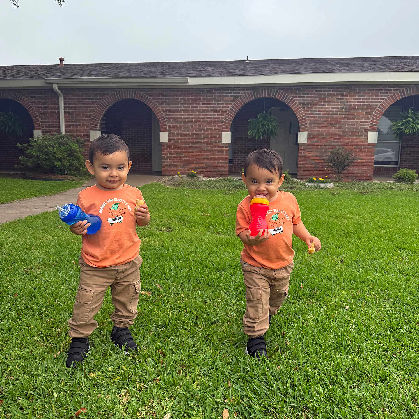 Mateo And Matias joined the competition — help win amazing prizes! child, toddler, grass, lawn, orange_shirt, khaki_pants, sippy_cup, snack, brick_building, arched_doorways, greenery, outdoor, two_children, matching_clothes, smiling, playful, cute, daylight, young_kids, casual_clothing