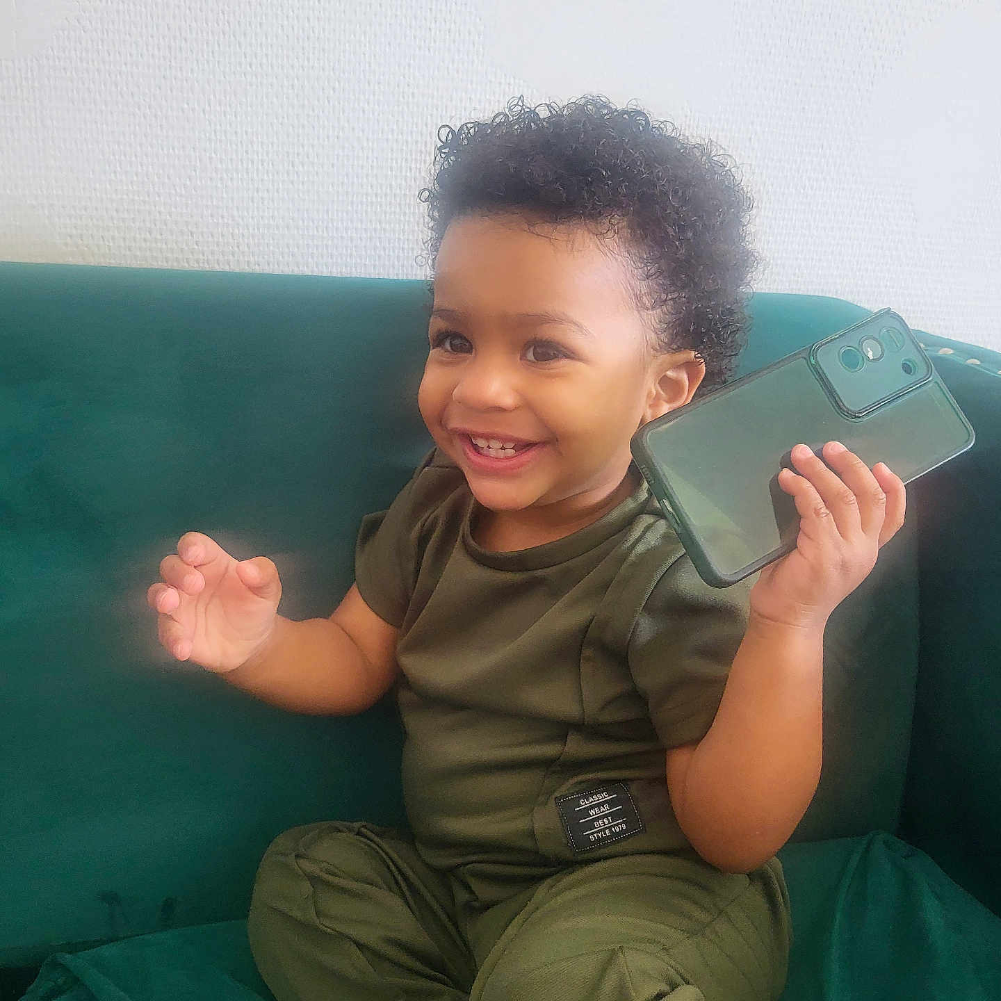 Jayann participe au concours pour gagner de l'argent avec cette photo : baby, casual, child, clothing, couch, curly_hair, cute, face, green_outfit, hand, happy, home, indoor, person, portrait, sitting, smartphone, smile, toddler, young_child