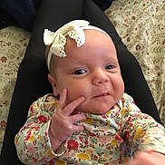 Paisley is registered to the contest to win money with this photo: baby, infant, headband, bow, floral_clothing, hand, face, person, lap, patterned_bedding, cozy, cute, smile, skin, eyes, fingers, indoors, portrait, child, young