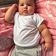 Valentina is registered to the contest to win money with this photo: baby, child, infant, face, arm, white_shirt, gray_pants, blanket, pink_blanket, soft_texture, cute, indoors, portrait, relaxed, curious, skin, human, laying_down, casual_clothing, young