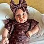 baby, infant, blue_eyes, pouty_face, headband, bow, brown_clothing, patterned_outfit, sitting, pillow, white_bedding, cute_expression, chubby_arms, fashion, portrait, indoor, child, soft_light, closeup, adorable