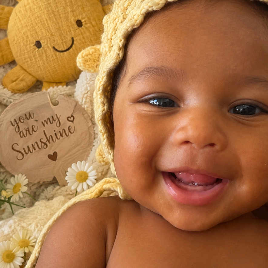 Jj is registered to the contest to win money with this photo: baby, infant, smiling, portrait, close_up, face, eyes, teeth, outdoor_style, sunhat, bonnet, plush_toy, sun, flowers, daisies, wooden_sign, blanket, happy, sitting, photography