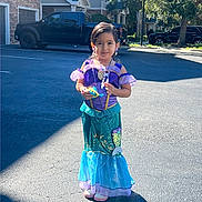 Elaine is registered to the contest to win money with this photo: child, girl, costume, mermaid, dress, toy, parking_lot, sunlight, smile, outdoor, residential_area, car, blue_sky, earrings, necklace, playful, shadow, standing, happy, accessories