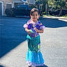 child, girl, costume, mermaid, dress, toy, parking_lot, sunlight, smile, outdoor, residential_area, car, blue_sky, earrings, necklace, playful, shadow, standing, happy, accessories