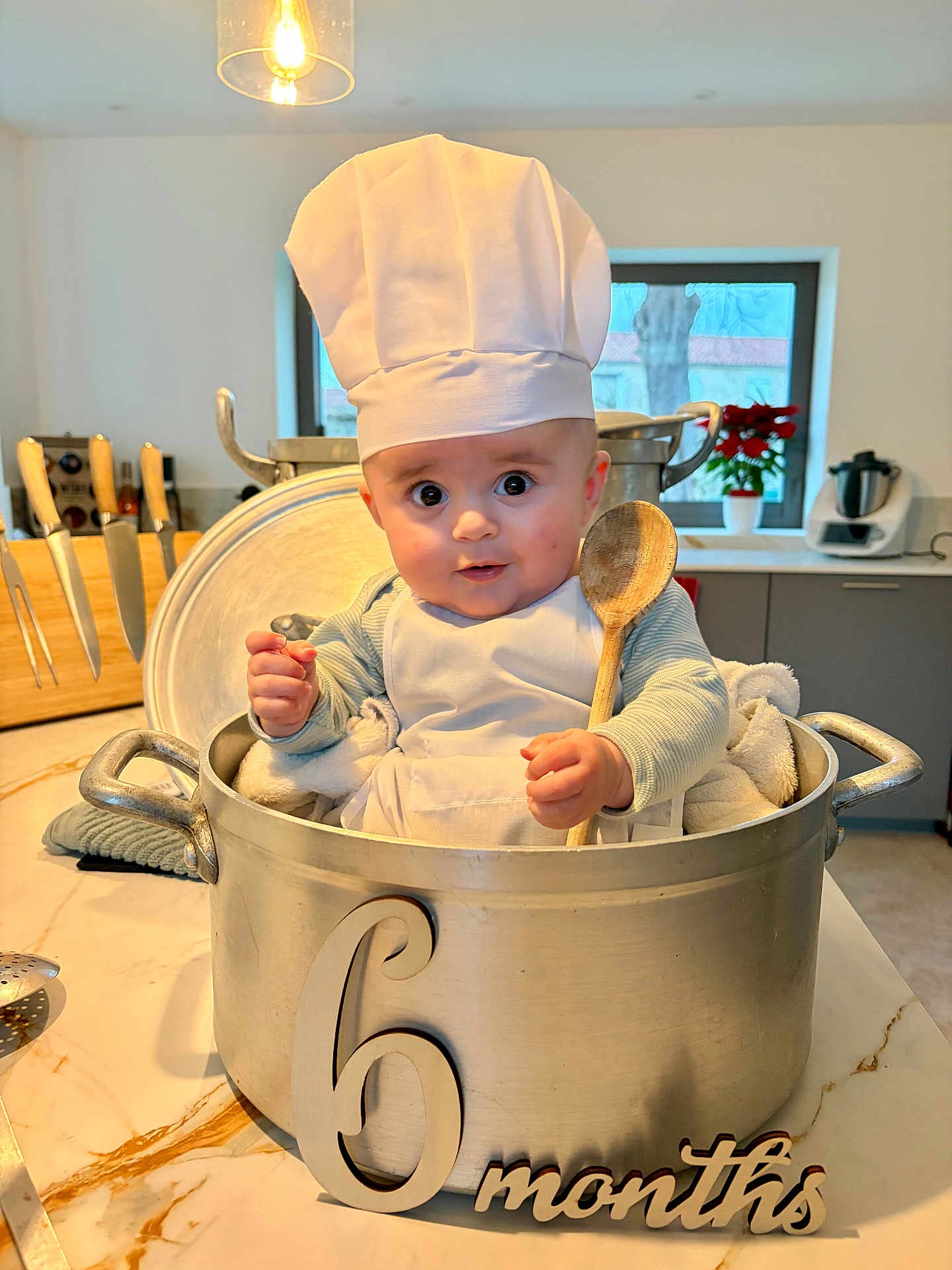 Lenny participe au concours pour gagner de l'argent avec cette photo : baby, infant, chef_hat, cooking_pot, wooden_spoon, kitchen, countertop, apron, cute, child, 6_months, utensils, window, plant, knife_block, expression, portrait, indoor, person, cooking