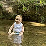 baby, child, water, stream, nature, outdoor, smile, happy, sitting, tank_top, diaper, shallow_water, greenery, sunlight, reflection, peaceful, summer, playful, young_child, natural_background