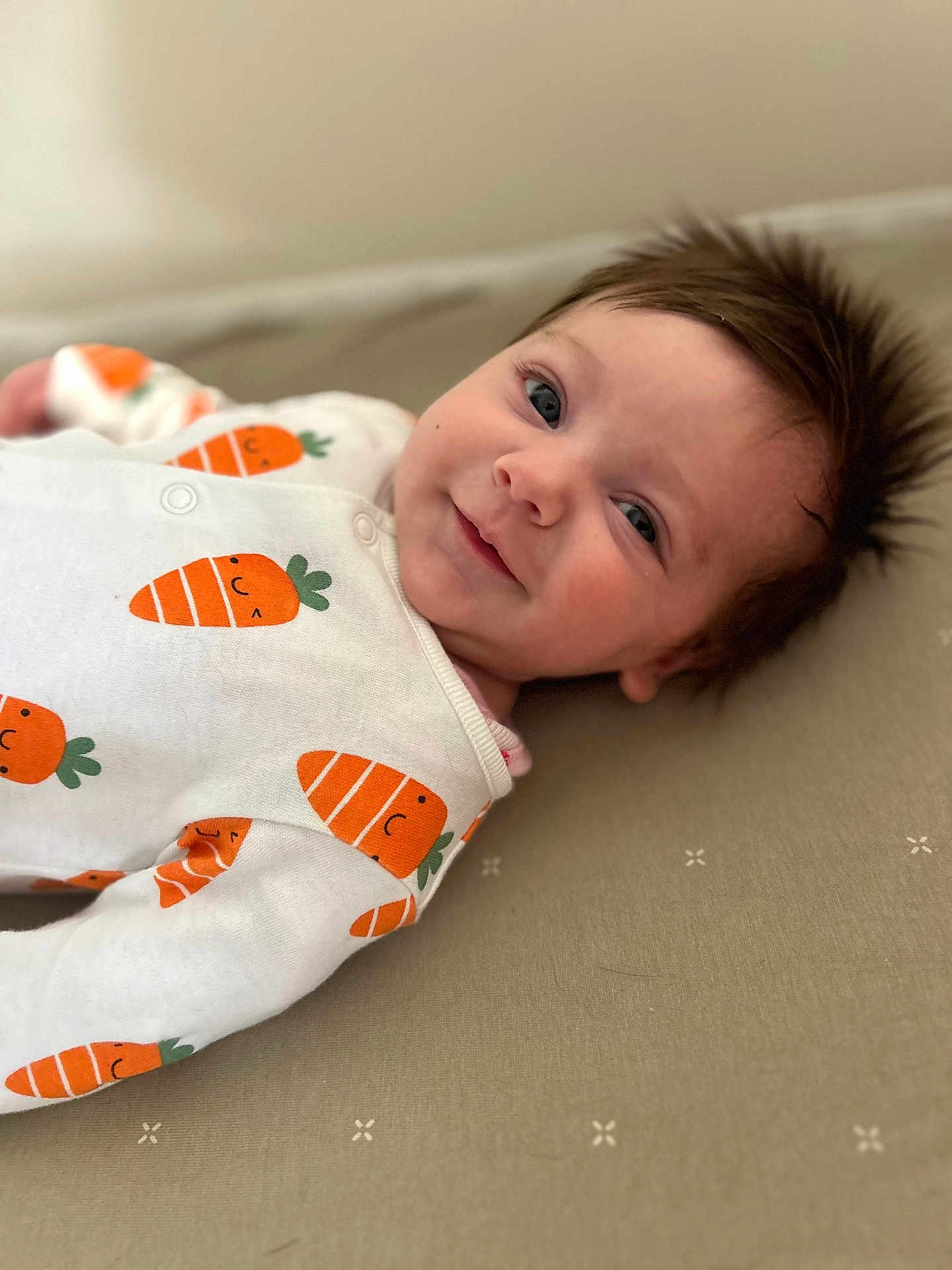 Eloise is registered to the contest to win money with this photo: baby, infant, child, lying_down, onesie, carrot_pattern, smile, face, blue_eyes, hair, soft_surface, cute, clothing, portrait, closeup, happy, skin, cozy, indoor, baby_garments