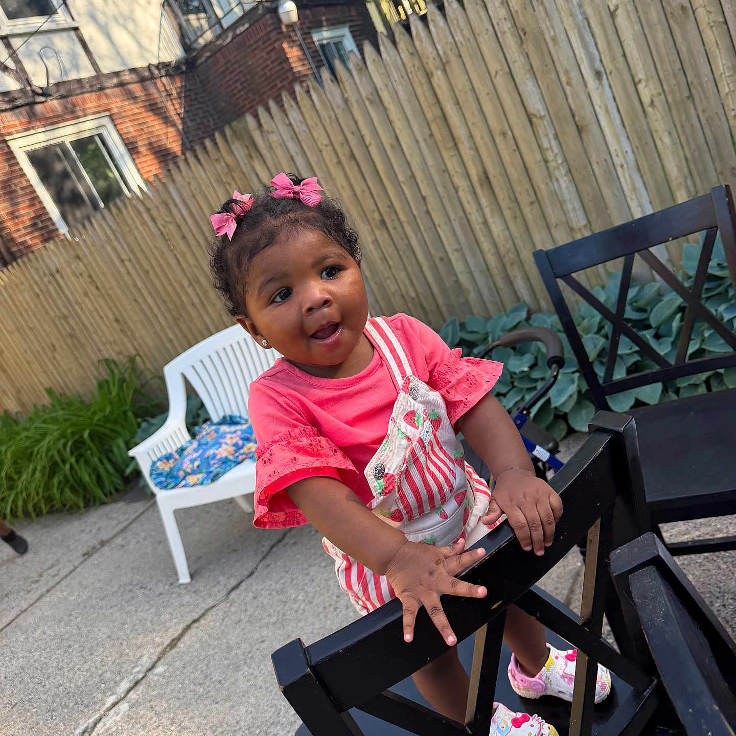 Saryha is registered to the contest to win money with this photo: toddler, child, pink_bows, outdoor, fence, patio, chair, smiling, playful, plants, concrete, house, clothing, footwear, person, daylight, garden, happy, wood, baby