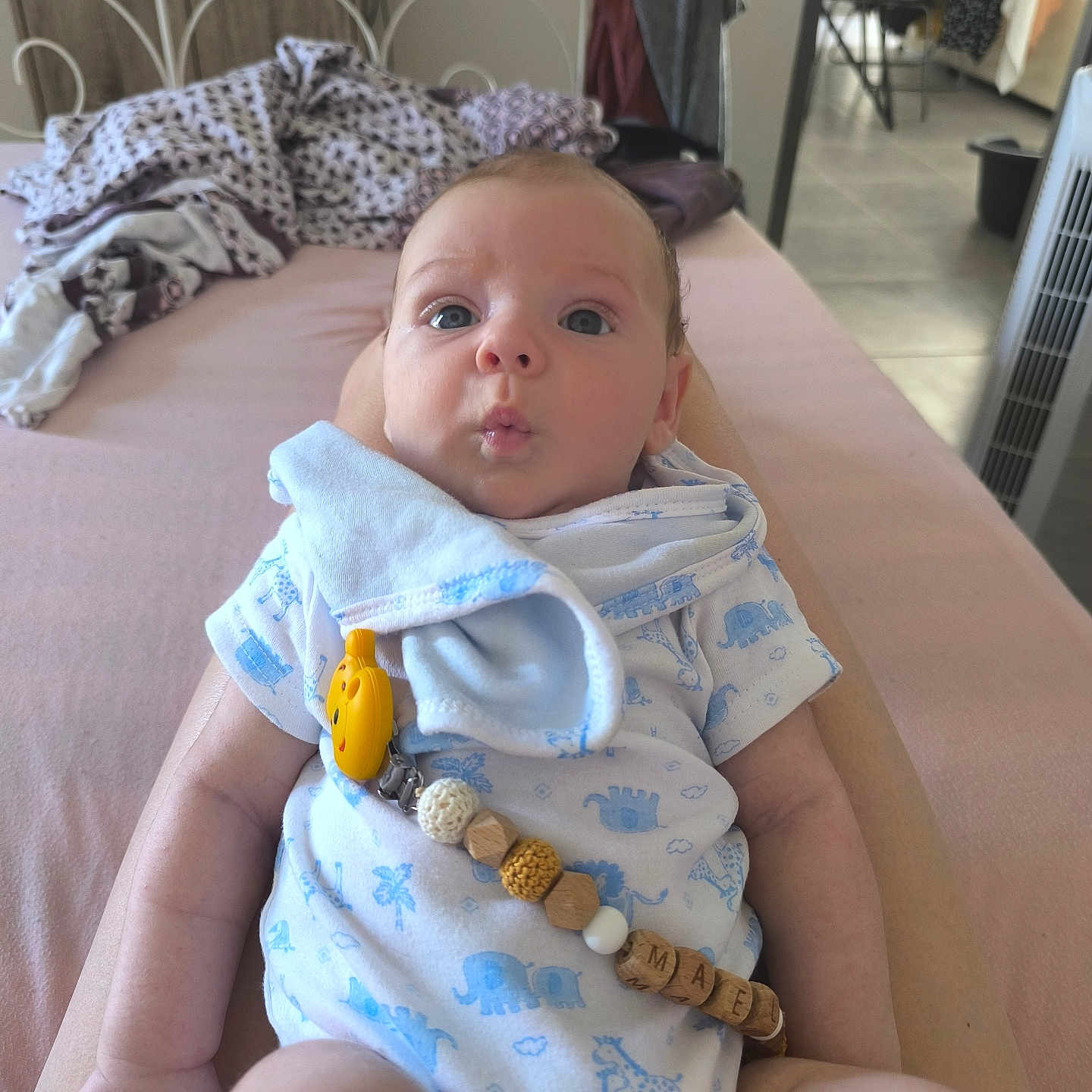 Maël participe au concours pour gagner de l'argent avec cette photo : animal_print, baby, bed, blanket, blue, clothing, curious, expression, face, furniture, hand, indoors, infant, legs, onesie, pacifier_clip, pink_bedspread, room, skin, soft