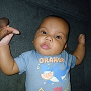 Michael is registered to the contest to win money with this photo: baby, infant, child, face, hand, tshirt, shark, orange, text, lying, fabric, dark_background, skin, cute, person, holding, finger, casual, clothing, portrait