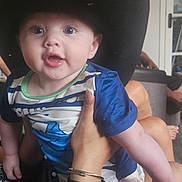 Jaxon joined the competition — help win amazing prizes! baby, hat, cowboy_hat, child, person, hand, indoor, blue_shirt, black_clothing, face, eyes, mouth, furniture, background, adult_hand, skin, expression, portrait, young_person, blurred_background