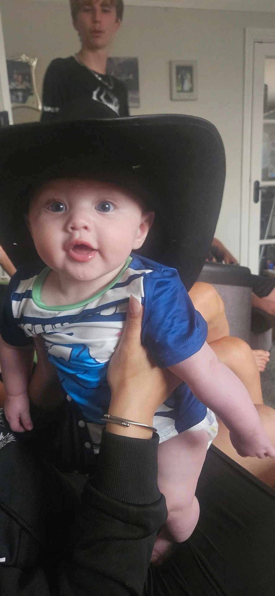 Jaxon joined the competition — help win amazing prizes! baby, hat, cowboy_hat, child, person, hand, indoor, blue_shirt, black_clothing, face, eyes, mouth, furniture, background, adult_hand, skin, expression, portrait, young_person, blurred_background