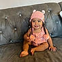 baby, child, smiling, pink_hat, pom_poms, knitted_hat, couch, gray_couch, indoors, happy, cute, sitting, infant, face, portrait, clothing, adorable, young_child, home, cozy