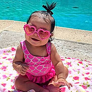 Chantelle is registered to the contest to win money with this photo: baby, child, sunglasses, heart_shaped, pink, swimsuit, pool, water, blanket, floral, toddler, outdoor, sunny, smiling, cute, hair_tie, sitting, barefoot, playful, summer