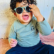 Aylan a rejoint le concours — aidez-le/la à gagner de superbes lots ! toddler, child, curly_hair, sunglasses, smiling, happy, sitting, blue_pants, long_sleeve_shirt, indoor, blanket, pillow, cute, fashion, portrait, playful, baby, casual_clothing, face, expression