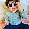 toddler, child, curly_hair, sunglasses, smiling, happy, sitting, blue_pants, long_sleeve_shirt, indoor, blanket, pillow, cute, fashion, portrait, playful, baby, casual_clothing, face, expression