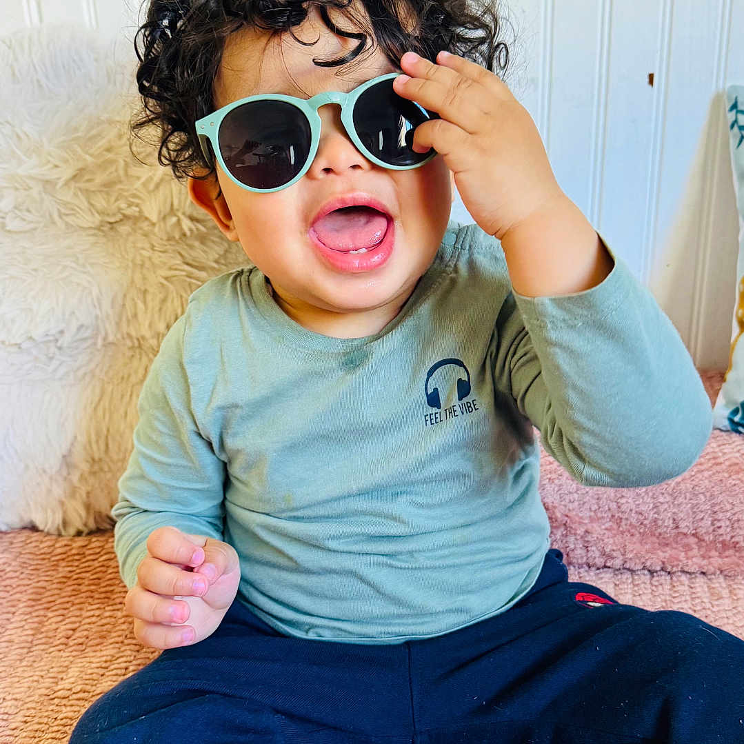 Aylan a rejoint le concours — aidez-le/la à gagner de superbes lots ! toddler, child, curly_hair, sunglasses, smiling, happy, sitting, blue_pants, long_sleeve_shirt, indoor, blanket, pillow, cute, fashion, portrait, playful, baby, casual_clothing, face, expression