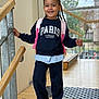 child, smiling, braided_hair, backpack, sweatshirt, indoor, railing, wooden_floor, window, natural_light, casual_clothing, happy, person, portrait, young, standing, fashion, footwear, school, ready
