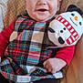 baby, smile, plush_toy, snowman, cushion, blanket, plaid_dress, red_sleeves, clothing, child, infant, happy, sitting, indoors, cute, soft_toy, face, person, cozy, portrait