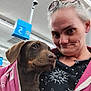 Cocoanuta is registered to the contest to win money with this photo: woman, dog, puppy, indoor, store, fluorescent_lights, jacket, pink, brown_dog, smile, portrait, person, clothing, eyewear, ceiling, sign, security_camera, shopping, closeup, expression