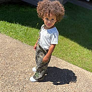 Kai is registered to the contest to win money with this photo: child, curly_hair, outdoor, sunlight, shadow, gravel_path, grass, white_tshirt, camo_overalls, shoes, standing, portrait, young_child, casual_clothing, daylight, nature, expression, curiosity, person, single_person