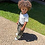 child, curly_hair, outdoor, sunlight, shadow, gravel_path, grass, white_tshirt, camo_overalls, shoes, standing, portrait, young_child, casual_clothing, daylight, nature, expression, curiosity, person, single_person
