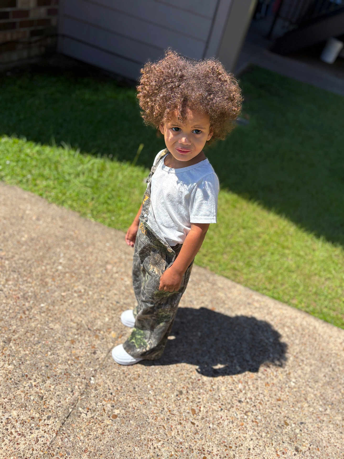 Kai is registered to the contest to win money with this photo: child, curly_hair, outdoor, sunlight, shadow, gravel_path, grass, white_tshirt, camo_overalls, shoes, standing, portrait, young_child, casual_clothing, daylight, nature, expression, curiosity, person, single_person