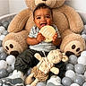 baby, child, smiling, plush_toy, teddy_bear, ball_pit, indoor, cute, holding, sign, sitting, clothing, white_sneakers, gray_pants, striped_shirt, happy, portrait, infant, playroom, soft_toys