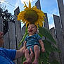 baby, infant, sunflower, flower, plant, smiling, happy, outdoor, fence, person, blue_sky, green_leaves, nature, holding, portrait, headwear_illusion, summer, daylight, closeup, human