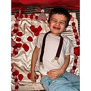 Joseph joined the competition — help win amazing prizes! child, boy, face, expression, white_shirt, jeans, suspenders_print, blanket, silk, felt_balls, heart_shape, red, pink, valentine_theme, indoor, playful, lying_down, casual_clothing, happy, cute